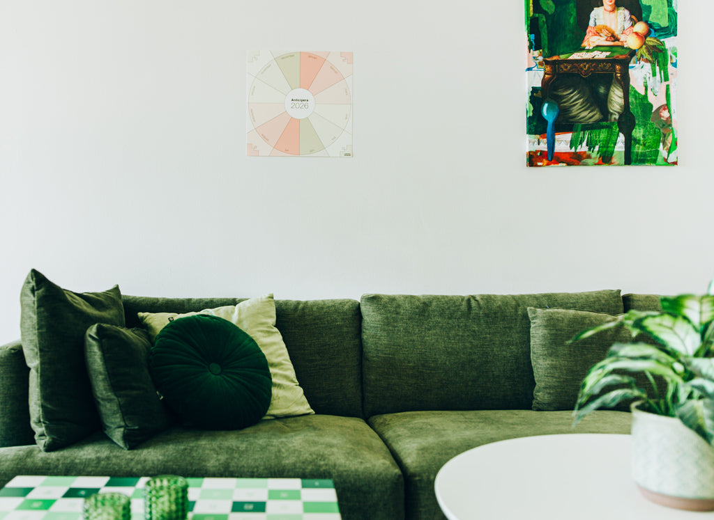 Anticipera väggplanerare i stilen Elin uppsatt på en vägg ovanför en grön soffa. Bredvid väggplaneraren hänger ett konstverk, i gröna färger.