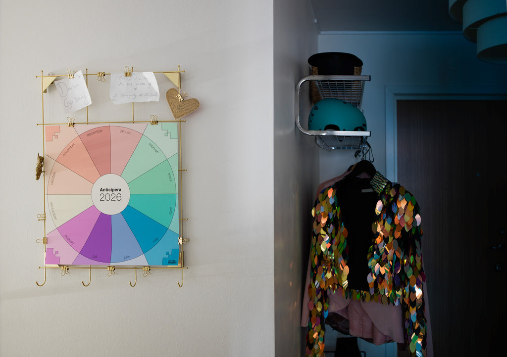 Anticipera väggplanerare i stilen Joan hänger på en anslagstavla i guld i en hall. I bakgrunden skymtar en paljettjacka, en cykelhjälm och en svart hatt. 