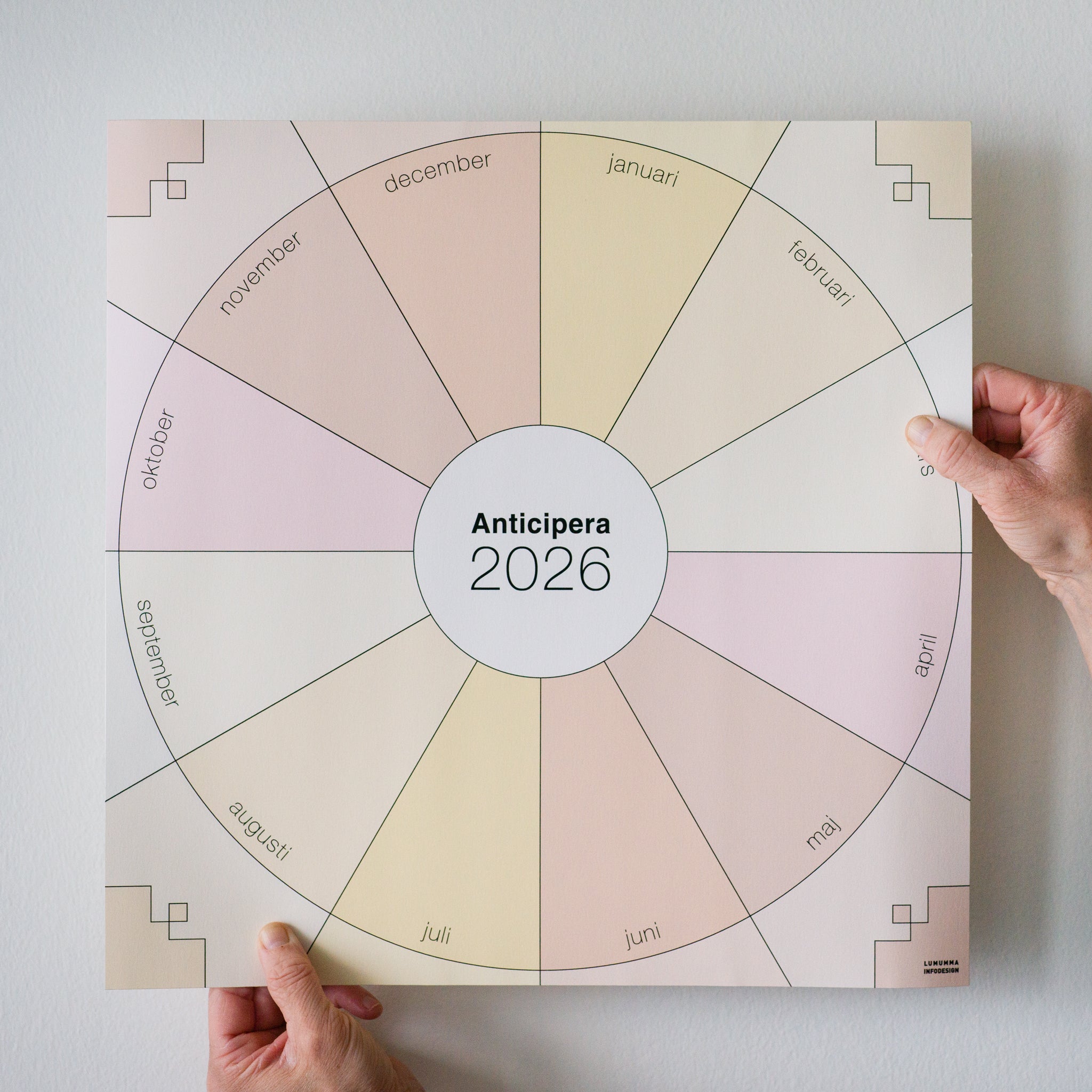 Ett par händer håller en Anticipera Väggplanerare årshjul 2026 i stilen Li med sex olika champagnefärger nära en vit vägg. I mitten står det Anticipera 2026.